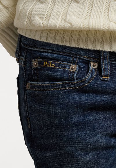 Vaqueros de denim azul oscuro con una cintura acanalada, presentando una etiqueta dorada "Polo" y herrajes plateados en los bolsillos. Textura sutil y detalles de costura.
