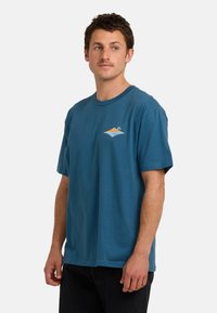 Mann mit kurzen Haaren, der ein blaues T-Shirt mit einem kleinen Grafik von einer tropischen Insel auf der Brust trägt, steht vor einem schlichten Hintergrund.