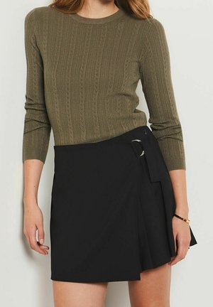 Kobieta w oliwkowo-zielonym, teksturowanym swetrze z dzianiny z rękawami do łokcia i czarnej spódnicy typu kopertowego z detalem z srebrną klamrą.