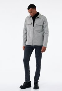 Chaqueta gris con cierre frontal y dos bolsillos de solapa en la parte delantera, textura suave, cuello con solapa, llevada sobre un suéter negro de cuello alto, combinada con jeans oscuros y zapatos negros.