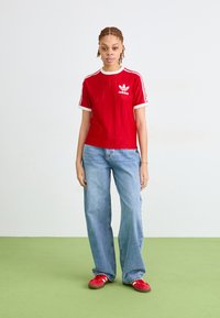 Rdeča kratka majica s kratkimi rokavi in vertikalnimi črtami, bela ovratnik in rokavi, vključuje logotip Adidas. Utečena z svetlo modrimi širokimi hlačami in rdečimi čevlji.