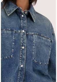 Blauwe denim overhemd met twee voorkeuringen, zilveren studs langs de rand van de zakken en parel-knopen. De stof heeft een gestructureerde afwerking.