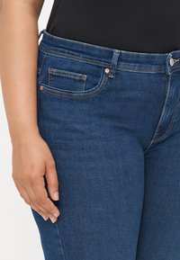 Dark blue jeans with a fitted design, featuring a front pocket, contrast stitching, and a metal button closure. Smooth denim texture.