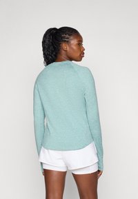 Haut de sport à manches longues en teal, fabriqué en tissu texturé avec un col rond. Il dispose d'un ourlet en courbe et de détails de ventilation discrets à l'arrière.