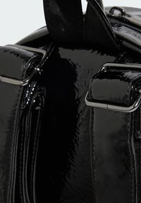 Dettaglio di una borsa in pelle verniciata nera con texture lucida, hardware in metallo e tracolle regolabili vicino a un compartimento con cerniera.