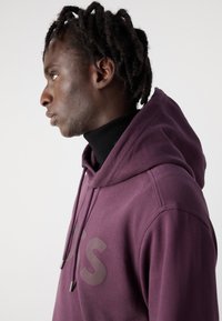 Bordeauxfarbener Kapuzenpullover mit einem großen grauen Aufdruck auf der Vorderseite, ausgestattet mit einer Kordelzugkapuze und einem gerippten Kragen. Glatte Stofftextur.