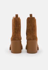 Botas de tobillo de ante marrón con tacón grueso, que presentan inserciones elásticas laterales y una lengüeta en la parte trasera. Superficie texturizada y suela rugosa.