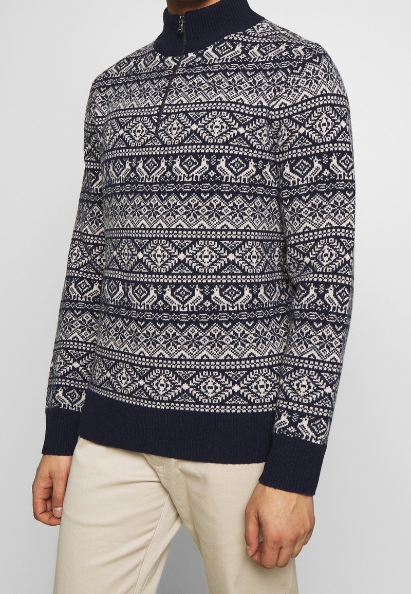Pull en maille à motif bleu marine et blanc avec col demi-fermeture éclair, porté avec un pantalon beige clair, ne montrant que la main et le torse.
