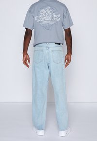 Grå grafisk t-shirt med texten "No Thanks" på ryggen, kombinerad med ljusblå högmidjade jeans. Modellen har ryggen mot kameran och bär vita sneakers.