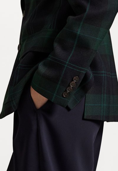 Osoba v tmavozelenom a čiernom károvanom blejzri s štyrmi gombíkmi na manžetách, rukou uvoľnene v vrecku námorníckych nohavíc.