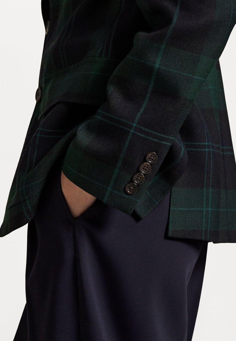 Persona che indossa un blazer a quadri verde scuro e nero con quattro bottoni sui polsini, mano casualmente nella tasca dei pantaloni blu.