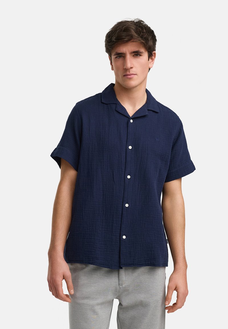 Camisa de manga corta azul marino hecha de tela texturizada, con cuello de punta y botones blancos, llevada con pantalones gris claro.