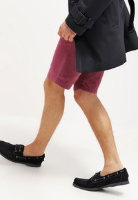 Maroon shorts with a simple design and a textured finish, paired with black slip-on shoes. The outfit includes a dark coat.