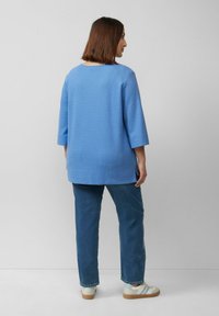 Lichtblauw gestructureerd bovenstuk met driekwartmouwen, gecombineerd met donkerblauwe jeans en witte sneakers met grijze accenten.