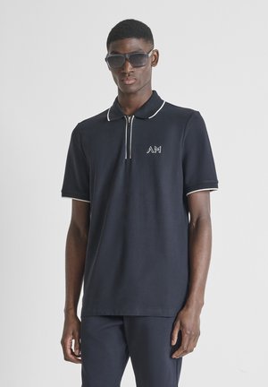 Jeune homme portant un polo bleu marine foncé avec bordure blanche, pantalon assorti et grandes lunettes de soleil noires sur un fond clair uni.