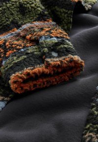 Gestrikte fleece stof in groen, zwart, bruin en oranje tinten, met een opgevouwen uiteinde dat zachte, donzige vezels toont tegen een gladde zwarte achtergrond.