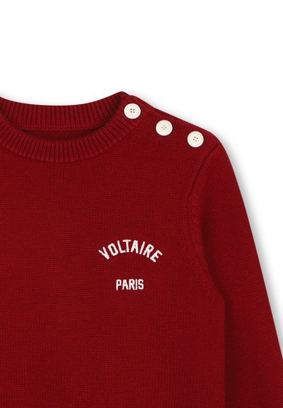 Κόκκινο πλεκτό πουλόβερ με λευκό κείμενο "Voltaire Paris" στο στήθος και τρία λευκά κουμπιά στον αριστερό ώμο κοντά στον ριμπ κολάρο.