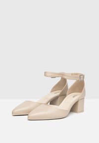 Scarpe beige in pelle con tacco a blocco, punta affusolata e cinturino alla caviglia con fibbia argento. Texture liscia, design minimalista.