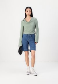 Blusa verde de manga larga con escote drapeado, combinada con pantalones cortos de mezclilla azul, con cintura ajustable y dobladillo enrollado. Zapatillas blancas y un bolso negro.