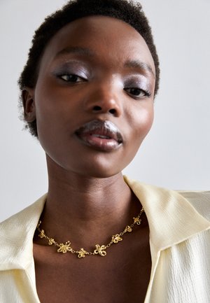 Collana in oro con motivi floreali, superficie liscia, indossata sopra una camicia gialla pallida. Il tono della pelle è marrone intenso. Sfondo semplice.