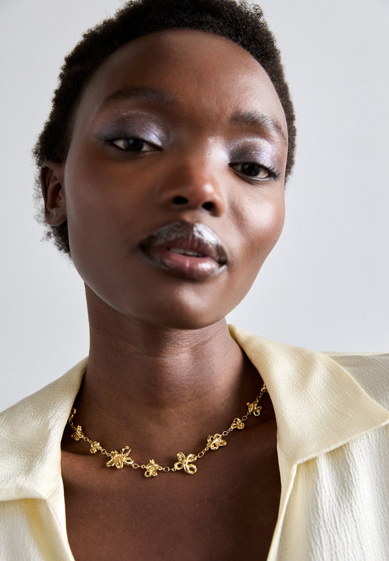 Gold chain necklace with floral motifs, smooth texture, worn over a pale yellow shirt. Skin tone is deep brown. Simple background.