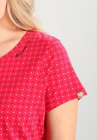 Camiseta roja con lunares que presenta mangas cortas, cuello redondo y texto bordado en el hombro. La etiqueta dorada en la manga incluye un logo.
