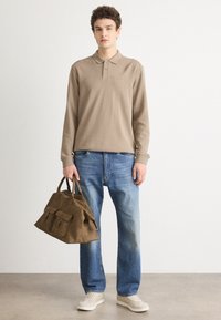 Beige langermet polo-shirt med krage, blå jeans og en brun semsket håndveske med to frontlommer. Lys grå bakgrunn.