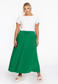 Groene maxi-jurk met een brede tailleband, gemaakt van zachte stof. Gecombineerd met een wit T-shirt met korte mouwen en lichtgekleurde platte schoenen.