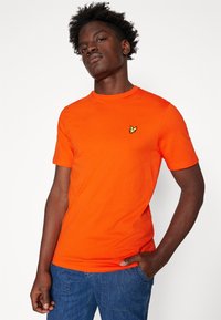 Leuchtend orangefarbenes T-Shirt aus Baumwolle mit einem Rundhalsausschnitt und einem kleinen gelben Vogel-Logo auf der linken Brust. Getragen mit blauen Jeans.