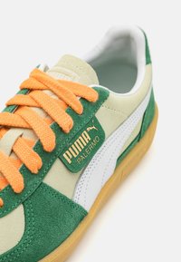 Sneaker verde in suede e pelle crema con lacci arancioni; presenta un'etichetta dorata "PUMA PALERMO" e una suola in gomma.