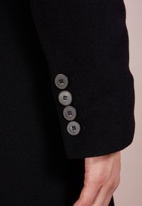 Zwarte wollen jas met een mouw die vier ronde, donkergrijze knopen met gegraveerde branding heeft. Gladde textuur, modern design, getailleerde pasvorm.