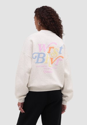 Frau mit lockigem Haar, die einen hellbeigen Pullover mit großem, buntem "Wrist Bhvr"-Schriftzug auf dem Rücken trägt, kombiniert mit einer schwarzen Hose.