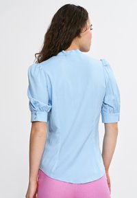 Jasnoniebieska bluzka z krótkim rękawem, z bufkowymi rękawami i detalami w postaci guziczków. Gładka tkanina z dopasowanym krojem i okrągłym dekoltem z tyłu.