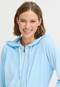 Světle modrá mikina na zip, měkká tkanina, s kapucí a stahovacími šňůrkami. Obsahuje stříbrný zip a volný střih. Usmívající se modelka v popředí.