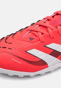 Botín de fútbol rojo con un upper sintético texturizado, acentos negros, rayas blancas y un logo prominente. Diseño con cordones y panel de malla.