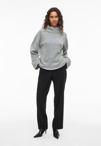 Grauer Strickpullover mit hohem Kragen und weiten Ärmeln, kombiniert mit schwarzen Hosen und schwarzen High Heels. Texturiertes Gewebe, lässige Passform.