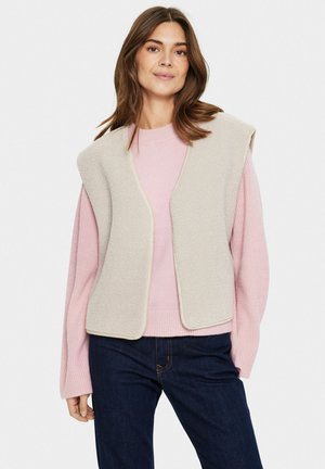 Beige fleeceväst med öppen front, rundade kanter och krämfärgade detaljer, lagd över en ljusrosa ribbad tröja, tillsammans med mörkblå jeans.