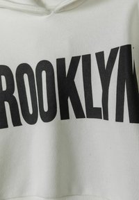 Vit huvtröja med stora svarta bokstäver som stavar "BROOKLYN". Materialet verkar mjukt med en strukturerad yta och ribbad nederkant.