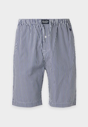 Homme torse nu debout pieds nus, portant un short rayé avec cordon, mains dans les poches, regardant vers la droite sur un fond clair uni.