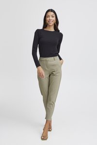 Kvinna som ler, klädd i svart långärmad topp, beige högmidjade byxor och gröna öppna klackskor, poserar med ena handen i fickan.