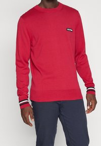 Pull rouge en tricot avec manches longues, col rond et un petit patch logo noir. Les poignets présentent des rayures rouge, blanc et bleu marine. Texture douce.