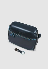 Bolso de cuero azul oscuro con un acabado suave, que presenta detalles de cremalleras en azul claro a contraste y un llavero desmontable. Forma rectangular.