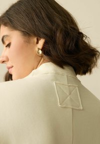 Cremfarvet beklædning med en tekstureret krave; har et broderet firkantet patch med krydset stingdetalje. Guld ørekam accent.