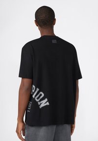 Man gezien van achteren in een losse zwarte T-shirt met grote grijze tekst "PION" en een grafisch ontwerp nabij de onderkant van de linker rug, gecombineerd met grijze broek.