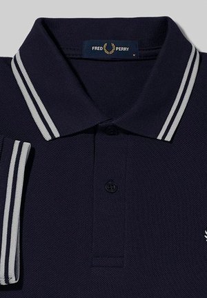 Polo Fred Perry azul marino con doble raya blanca en el cuello y los bordes de las mangas, con tapeta de botones y el logo de la corona de laurel.