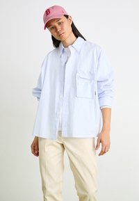 Cămașă oversized în dungi albastre deschise, cu mâneci lungi și un buzunar pe piept, asortată cu pantaloni bej și o șapcă roz cu un logo.