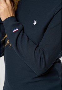 Sötétkék, hosszú ujjú póló texturált anyagból, fehér hímzett logóval a mellkasán és egy kis háromszínű díszítéssel az ujjon.
