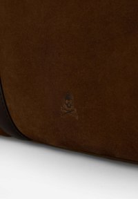 Bolso de ante marrón con un sutil logo de calavera y huesos cruzados. Superficie texturada con acentos de cuero oscuro en contraste. Forma rectangular.