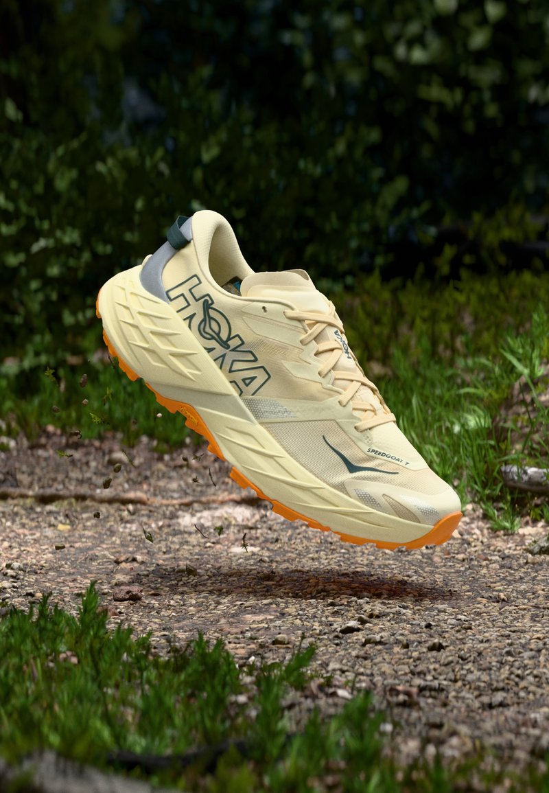 Lys gul løbesko med et tekstureret overmateriale, grå accenter og en orange ydersål, der har et polstret design for bedre greb.