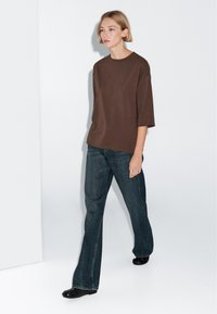 Woman with short blonde hair walking, wearing a loose brown top with elbow-length sleeves, dark blue wide-leg jeans, and black shoes.